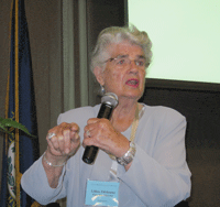 An elderly woman with gray hair, wearing glasses and a light blue outfit, speaks into a microphone while gesturing with her hand during a presentation. A name badge hangs from her neck.