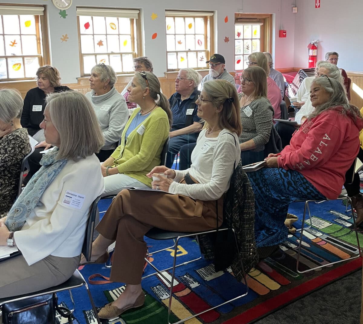 Adults sit attentively in a classroom with colorful windows and a bright rug, some holding notebooks and wearing name tags, as they listen to a speaker out of view.