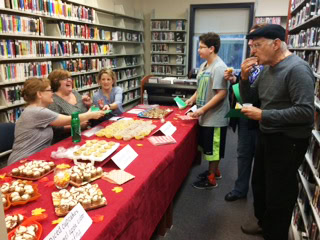 A group of people sample baked goods at a table covered with treats in a library. Some are handing out or eating food, while shelves of books line the background.
