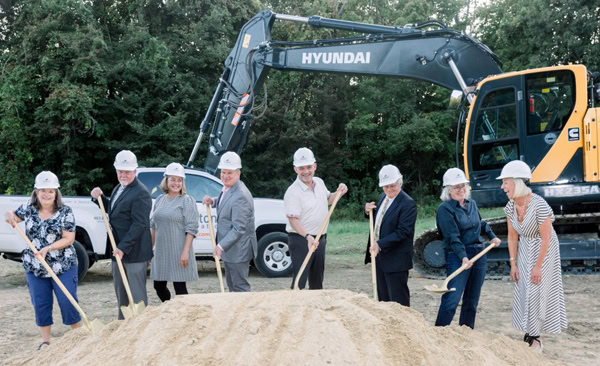 Eight people in business attire and hard hats stand in front of a pile of dirt with shovels, smiling for a groundbreaking ceremony. A Hyundai excavator and trees are visible in the background.