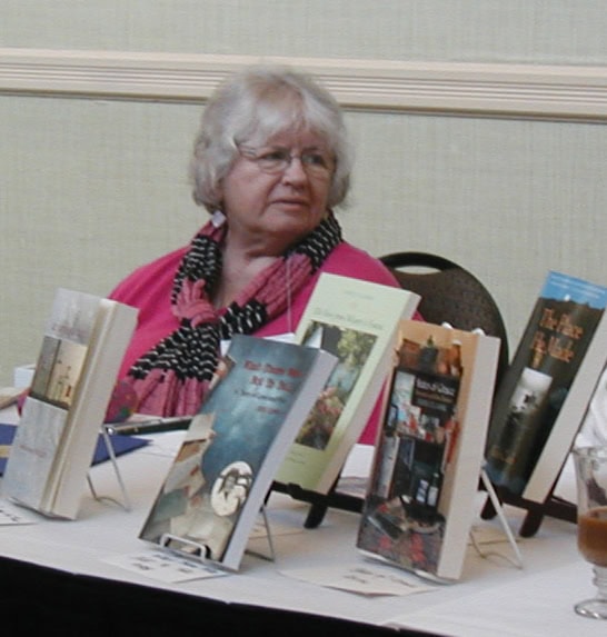 An older woman with gray hair and glasses sits at a table displaying several books on stands. She wears a pink top and a scarf, and looks to the side. The background is a light-colored wall.