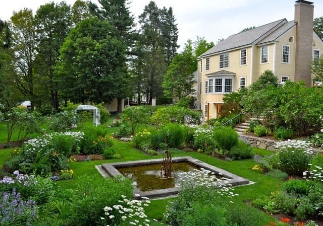 A lush garden with blooming flowers and a square reflecting pond sits in front of a large beige house with many windows, surrounded by tall trees and greenery.
