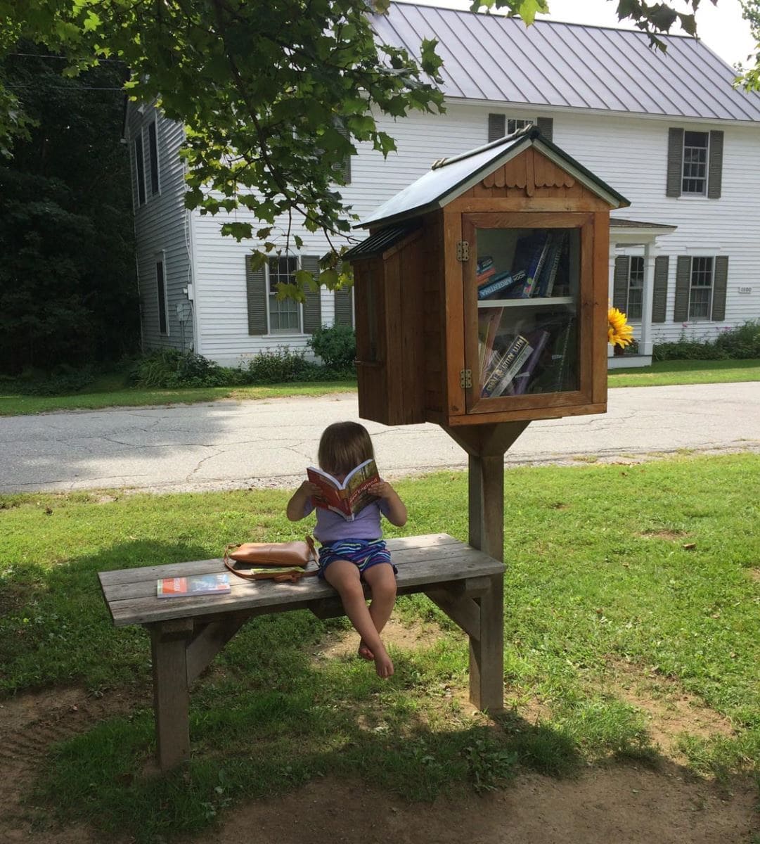 A young girl sits on a wooden bench reading a book beside a small outdoor community library box on a grassy lawn, with a white house and a quiet street in the background.