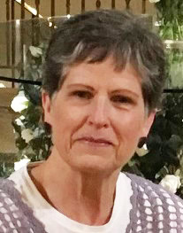 A woman with short gray hair and a serious expression wears a light-colored top and sits indoors, with greenery and white flowers in the blurry background.