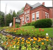 A red-brick building with a triangular roof sits behind a garden full of colorful flowers and greenery, with a sign and a path leading up the landscaped slope.