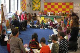 A classroom with children sitting on a blue carpet, reading books. Several adults sit at the front, also holding books. Colorful artwork and educational materials decorate the walls.
