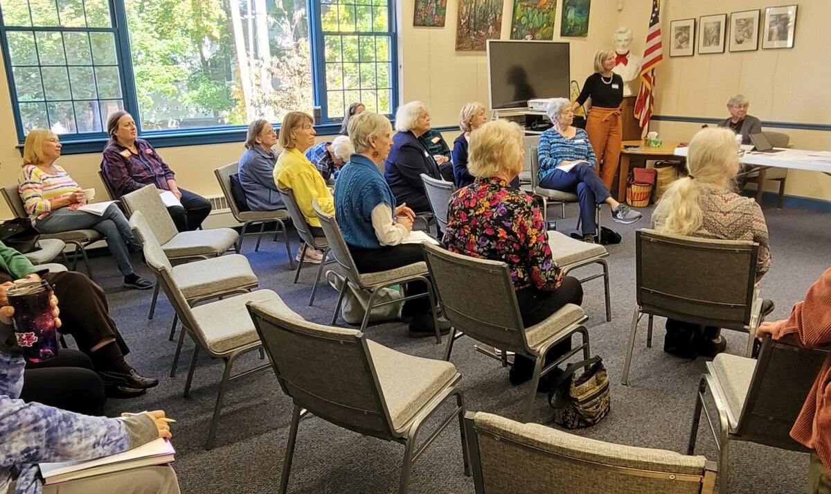 A group of older adults sit spaced out on gray chairs in a community room, listening to a standing woman near an American flag. Art hangs on the walls, and sunlight streams through large windows.