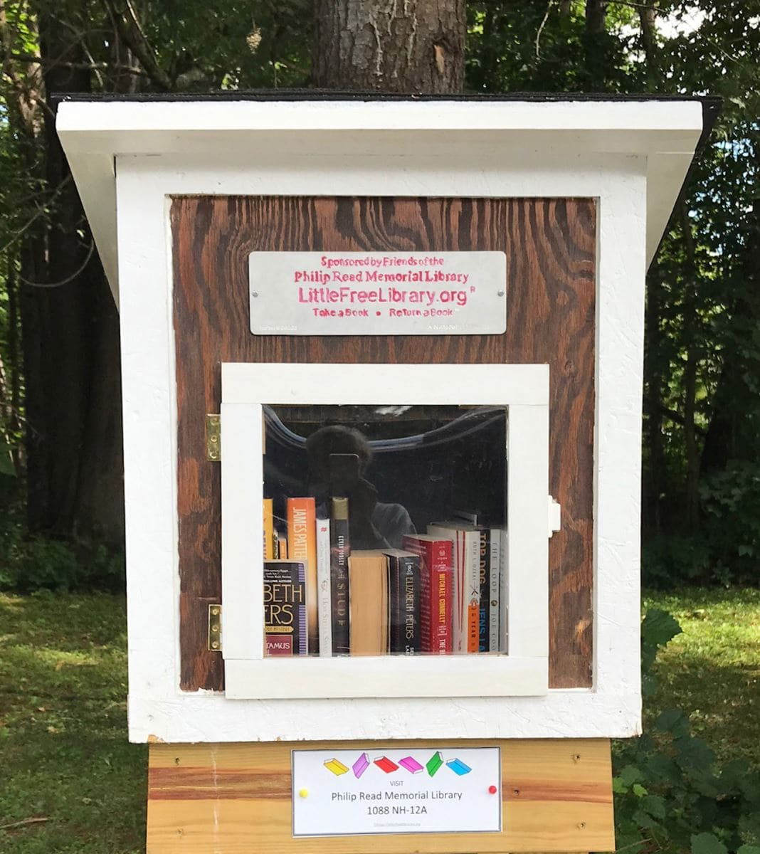 A small outdoor wooden box labeled Little Free Library holds several books. The box has a glass-front door and sits against a backdrop of trees and grass. A sign promotes the Philip Read Memorial Library.